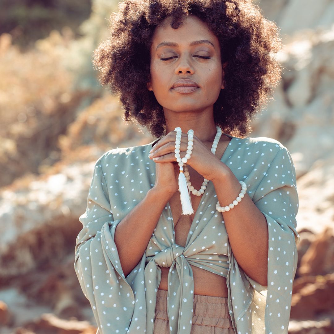 Woman standing outside with eyes closed wearing Ambarya New Beginnings - Moonstone Mala Bead Set. The mala bead necklace is draped over her fingers 